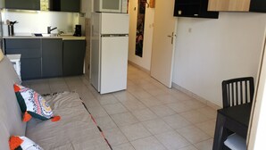 Cocina compacta con microondas y refrigerador, sofá con cojines estampados, y un comedor con una silla.