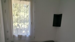 Un dormitorio pequeño y ordenado con una cama individual, una mesita de noche con lámpara y una ventana con cortinas de gasa.