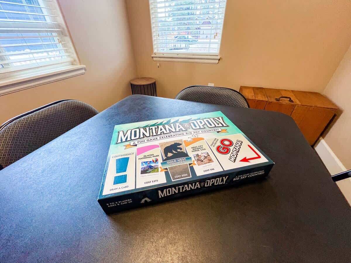 A game box titled 'Montana Opoly' on a table in a room with two windows and a chair.