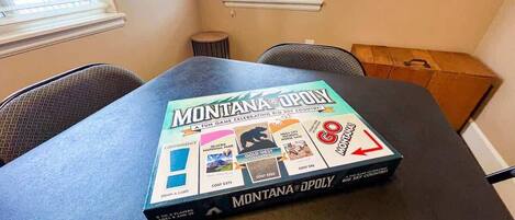 A game box titled 'Montana Opoly' on a table in a room with two windows and a chair.