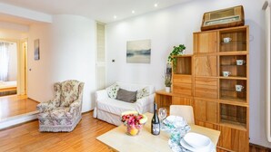 A living room with a wooden cabinet, a floral armchair, a white sofa, and a dining table with a fruit bowl and wine bottle. - GuestHost - Ponte Solferino Bright & Cozy APT (Pisa)