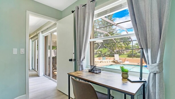 A room with a desk, chair, and a view of a swimming pool through a large window with curtains. - Casa Boca - Perfect Florida Getaway with Large Heated Saltwater Pool (Boca Raton)