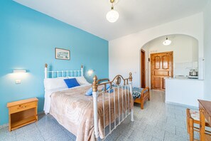 A bedroom with a bed, bedside tables, a wooden bench, a kitchen area with a sink and cabinets, and a door leading to another room.