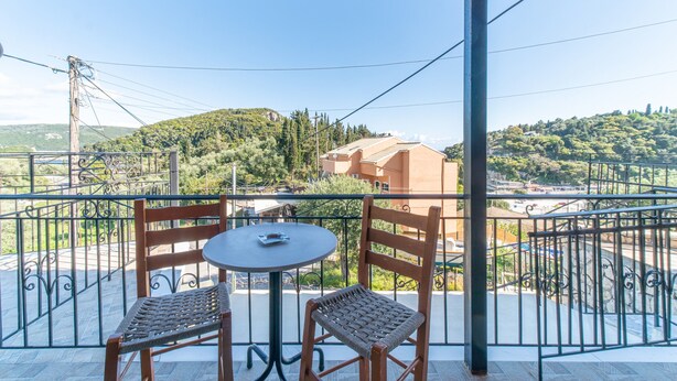 Ein Balkon mit zwei Korbstühlen und einem kleinen runden Tisch, mit Blick auf eine hügelige Landschaft mit Gebäuden.