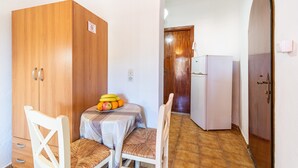 Ein kleines Zimmer mit einem Holzschrank, einem Tisch mit Früchten, einem Stuhl und einem weißen Kühlschrank.