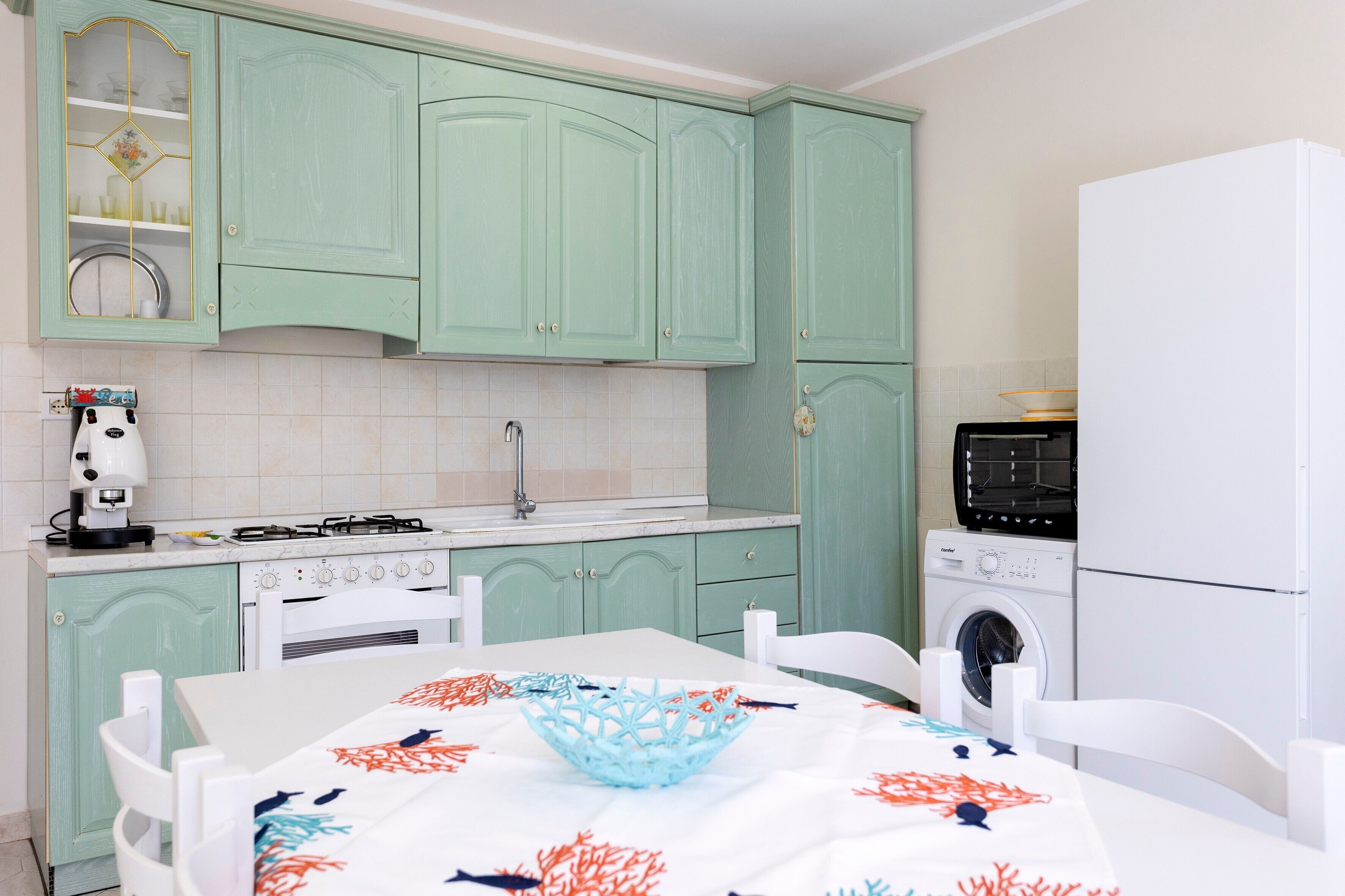 Una cucina con mobili color petrolio, un tavolo bianco con tovaglia a fiori e una lavatrice.