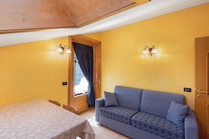 A room with a bed, a sofa, two wall-mounted lights, a wooden door, and a window with curtains.