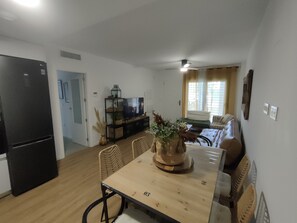 Un comedor con una mesa de madera, sillas, un refrigerador negro, un televisor sobre una estantería y una ventana con cortinas.