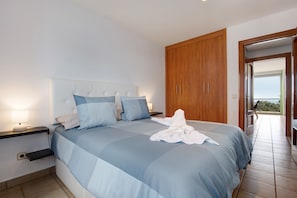 Une chambre à coucher avec un lit, des tables de chevet, une armoire en bois et une vue sur l’extérieur par une porte ouverte.