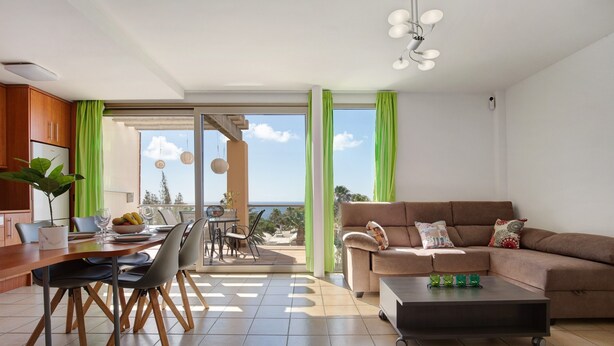En moderne stue med sofa, sofabord, spiseplads og en balkon med udsigt.