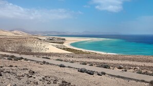 Et kystlandskab med en asfalteret sti, en sandstrand og et klart, blåt hav.