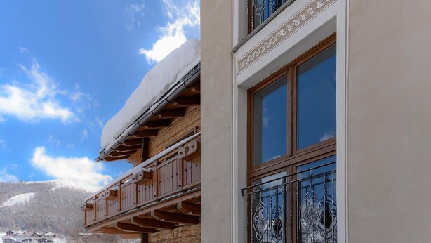 A log cabin with a balcony, a window with decorative ironwork, and a snowy landscape in the background.