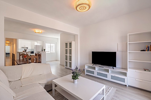A modern living room with a white sofa, a coffee table, a TV on a white stand, and a bookshelf.