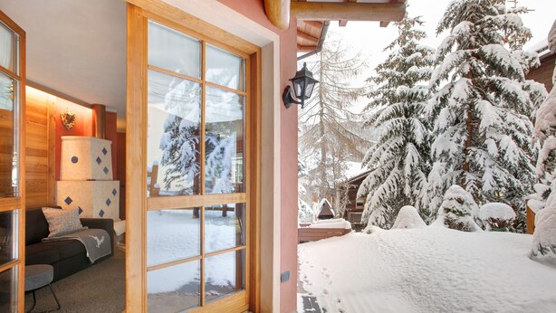 Una habitación con estructura de madera, puerta de vidrio que da al exterior, sofá con cojines y una mesita con lámpara.