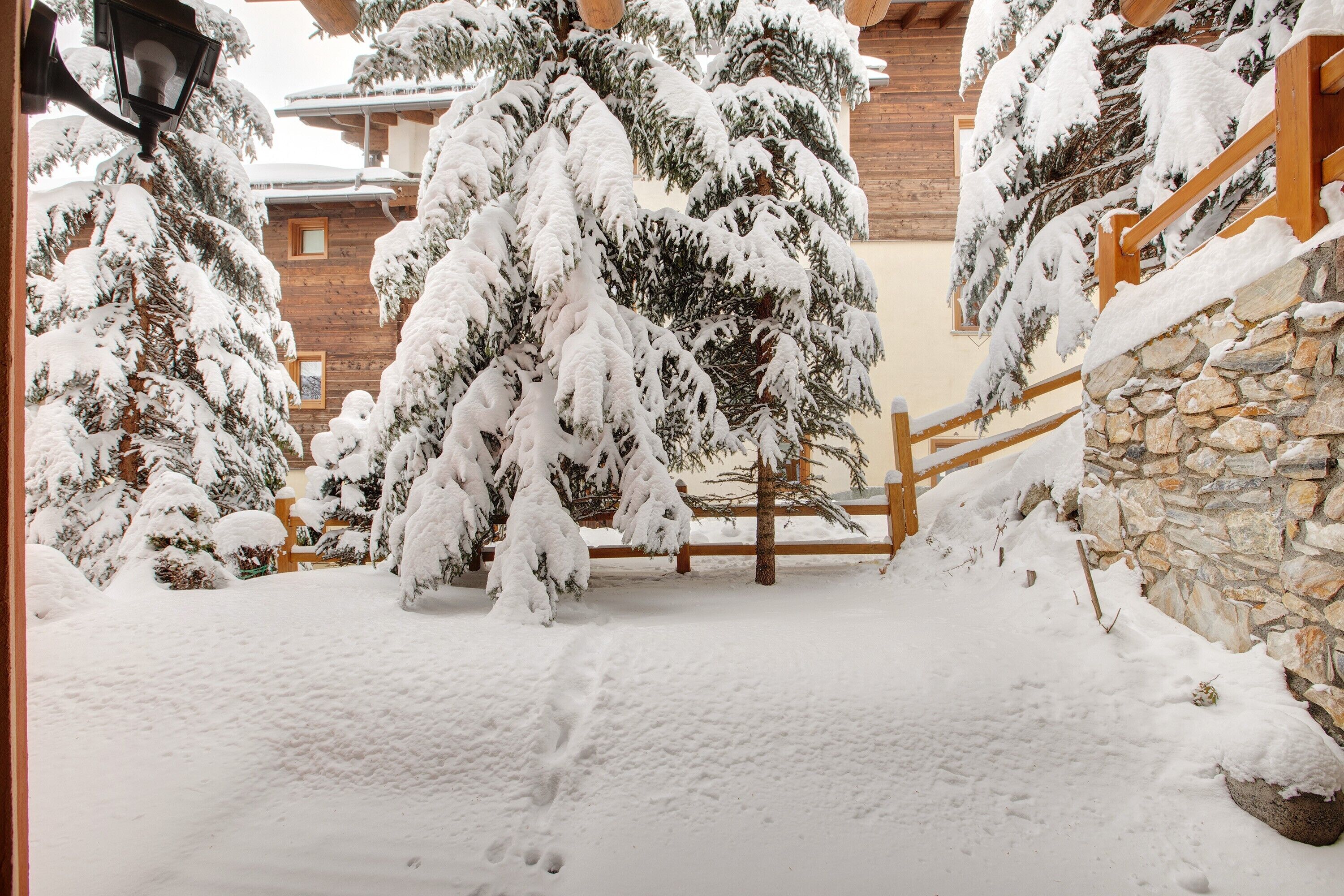Un’area esterna innevata con recinzioni in legno, mura in pietra e alberi carichi di neve.
