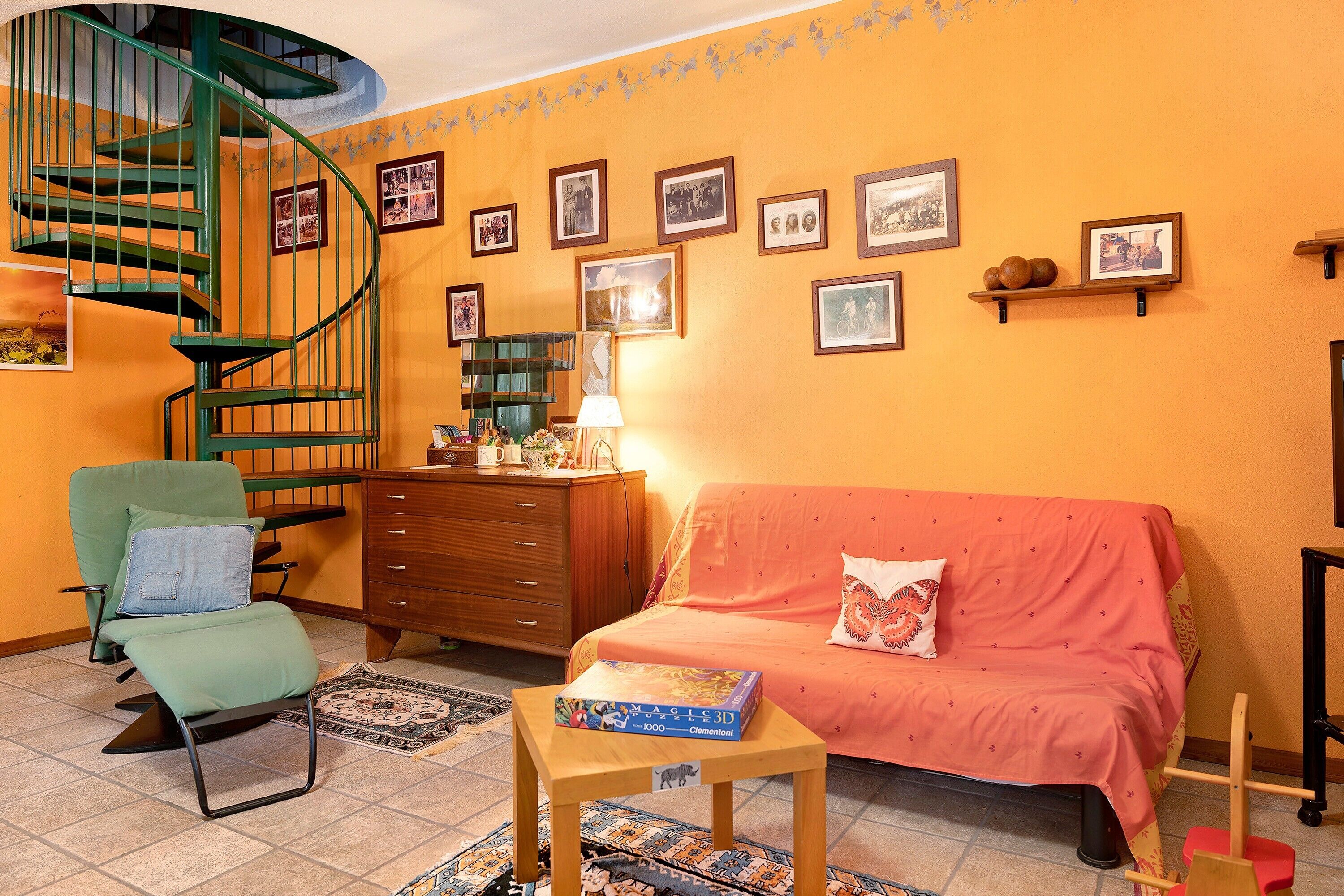 Een kamer met een spiltrap, een groene fauteuil, een houten dressoir, een rode bank en een kleine tafel met een spel.