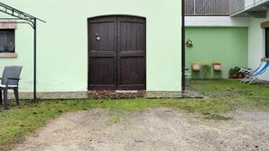 A building with a large wooden door, a small outdoor seating area with chairs, and a grassy courtyard.