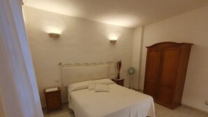 A bedroom with a bed, bedside tables, a nightstand, a wooden wardrobe, and a standing fan.