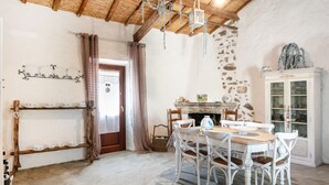 A rustic room with a stone wall, wooden ceiling, a fireplace, a dining table with chairs, and a cabinet with glass doors. - Holiday Home 'Stazzu Saiacciu' with Terrace (Palau)