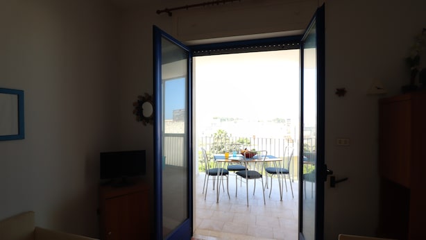 Een open deur die leidt naar een balkon met een tafel en stoelen, een televisie op een standaard en een klok aan de muur.