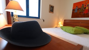 Een kamer met een bed, een lamp, een hoed op een tafel en een schilderij aan de muur.