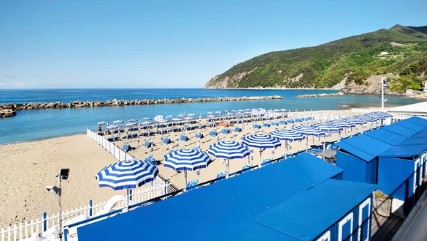 Una playa con hileras de sombrillas azules y sillas reclinables, un litoral rocoso y una colina al fondo.