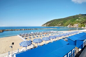 Una spiaggia con file di ombrelloni blu e lettini, una costa rocciosa e una collina sullo sfondo.
