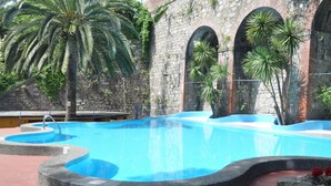 Una piscina amb mur de pedra i obertures apuntades, envoltada de palmeres i un paviment de rajoles vermelles.