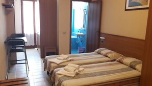 Habitació amb dos llits, un escriptori, una cadira, i un bany visible a través d'una porta oberta, més un finestral amb cortines.