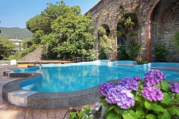 A swimming pool with a stone edge, surrounded by greenery and a stone building with arches.