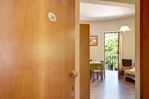 A wooden door with a number plate, a dining area with chairs and a table, a bed with a TV, and a balcony with a view.