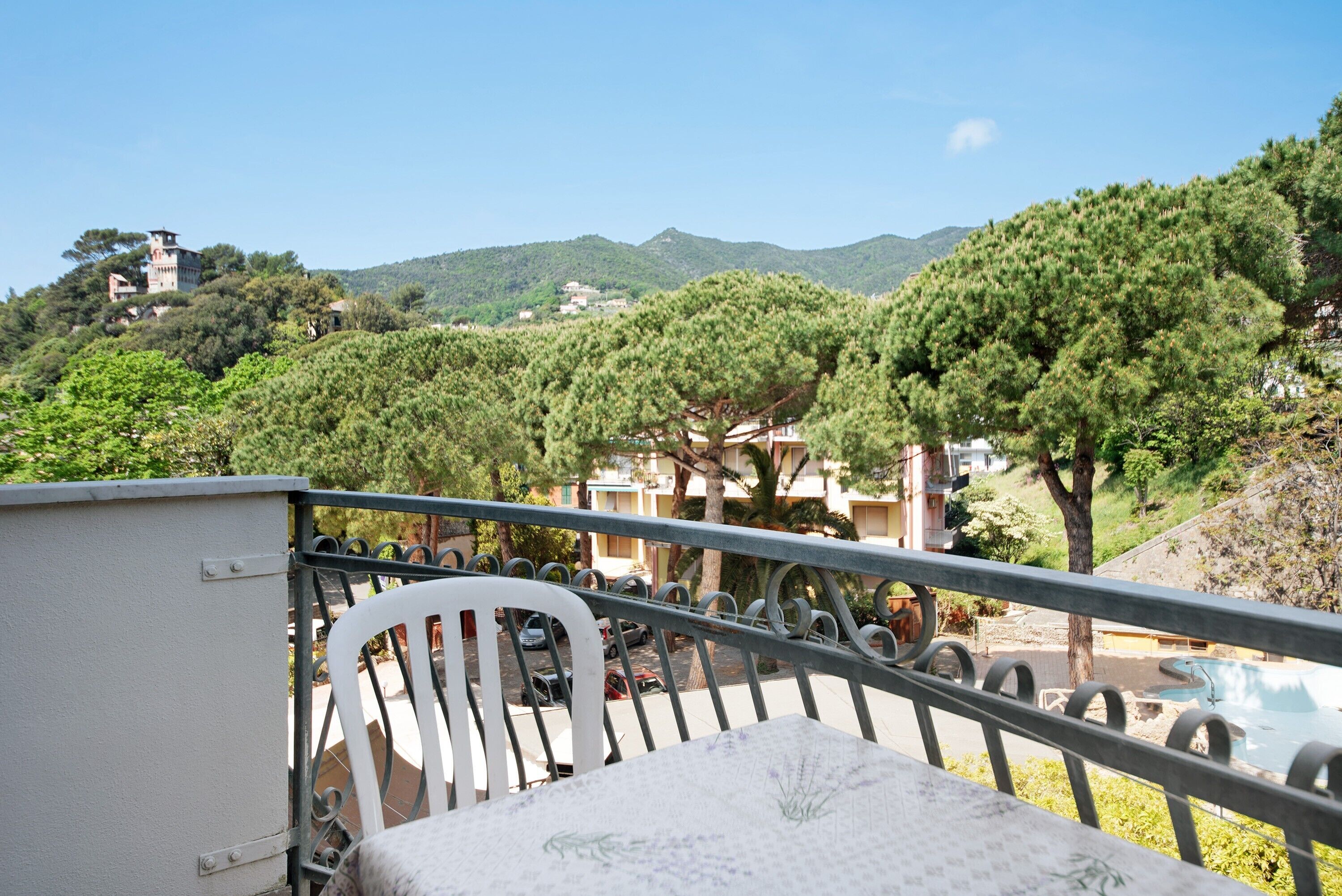 A balcony with a white chair, a table, and a view of a pool and buildings in the distance.