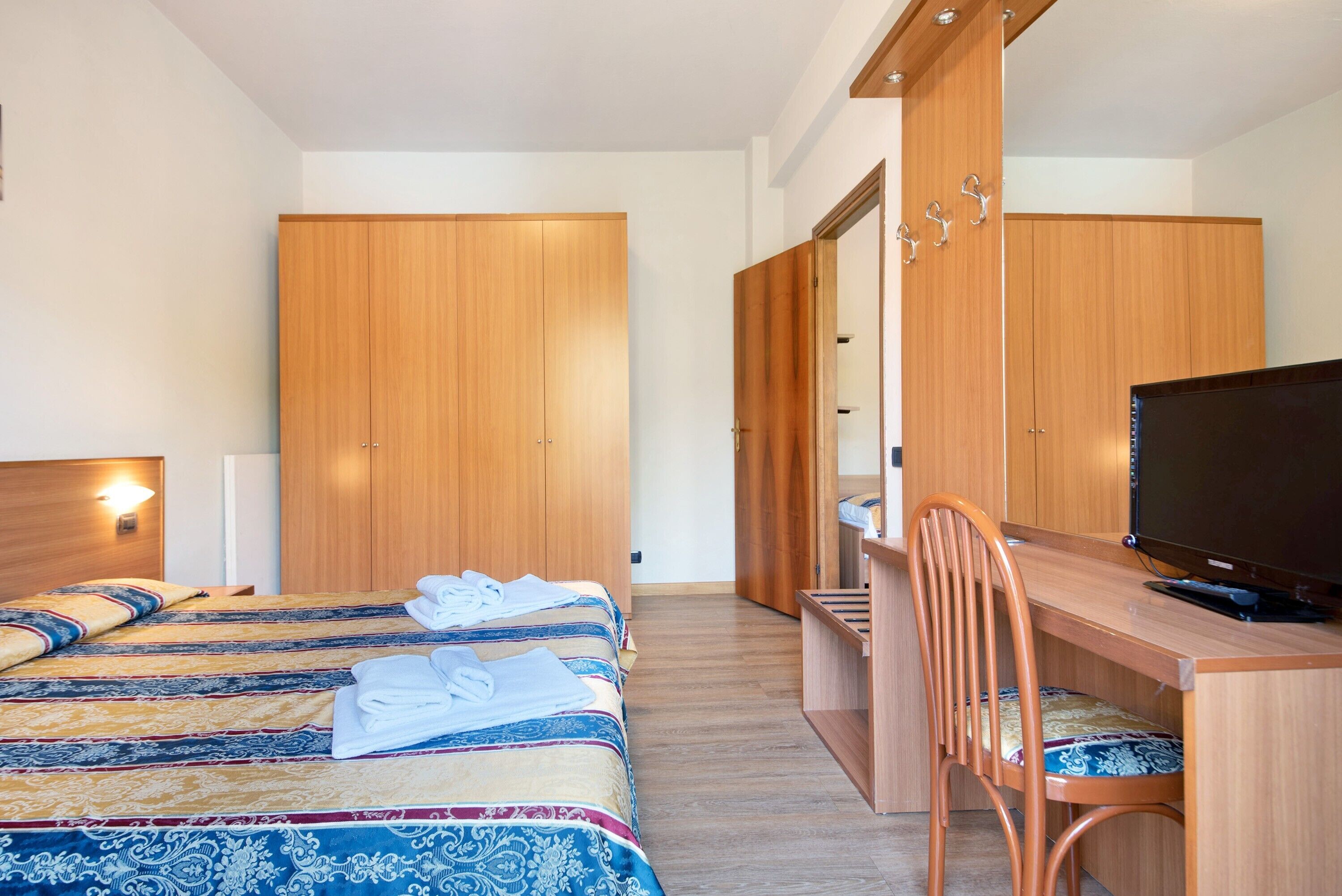 A hotel room with a bed, a desk with a chair, a television, and wooden wardrobes.