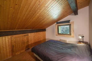 A bedroom with a sloped ceiling, wooden walls, a window with a view of trees, a bed, and a bedside lamp.