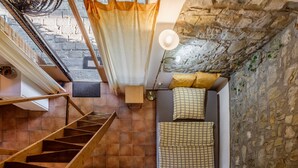 A room with a stone wall, a bed with checkered bedding, a wooden staircase, and a hanging light fixture.