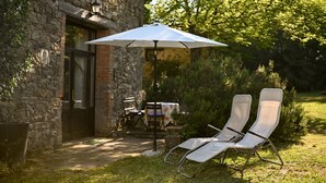 Un bâtiment en pierre doté d’une terrasse, de deux fauteuils de jardin, d’une table avec des chaises et d’un parasol.