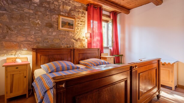 Ein Schlafzimmer mit einem hölzernen Bett, einer Steinwand und einem Fenster mit roten Vorhängen.