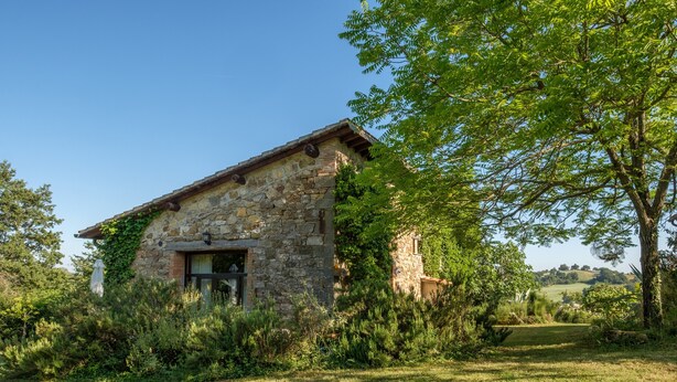 Un bâtiment en pierre doté d’un toit en pente, entouré de verdure et surplombé d’un ciel dégagé.