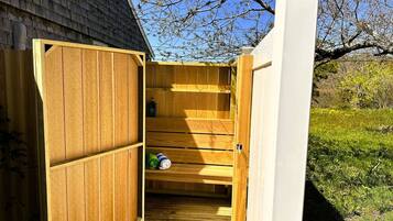 A wooden sauna with a bench and shelves, set against a house with a white exterior and a tree in the background.