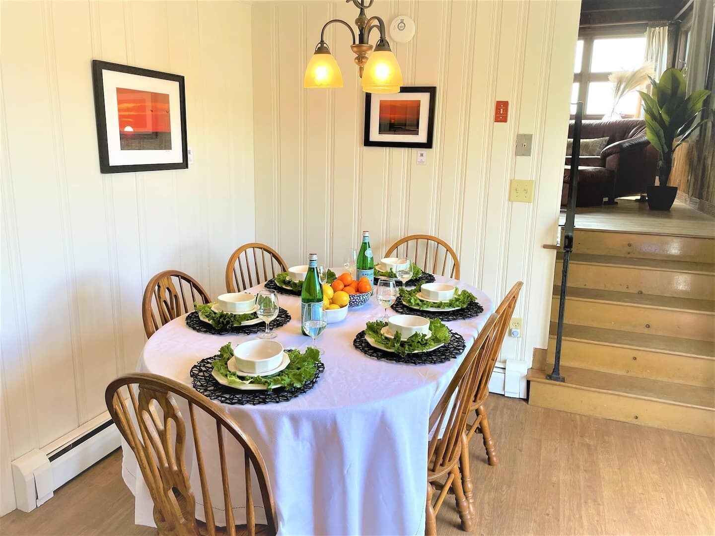 Mesa de comedor preparada para seis personas, con mantel blanco, individuales negros y hojas verdes. Alrededor de la mesa hay sillas, una botella de vino y un cuenco de fruta.