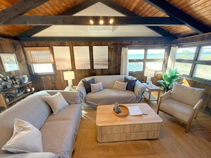 Una habitación acogedora con techo de madera, un sofá a rayas, un sofá color beige, una mesa de centro de madera y vistas al exterior a través de amplias ventanas.