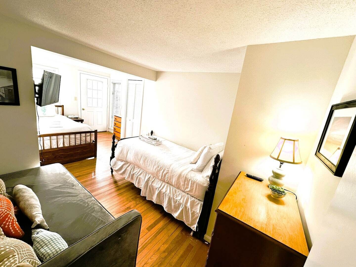 A small, well-lit room with a bed, a wooden nightstand, a lamp, and a couch.
