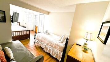A small, well-lit room with a bed, a wooden nightstand, a lamp, and a couch.