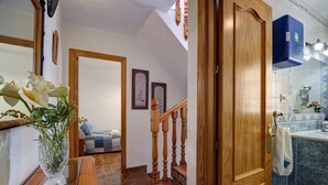 Un couloir avec un escalier en bois, un vase rempli de fleurs et une salle de bain aux carreaux bleus.