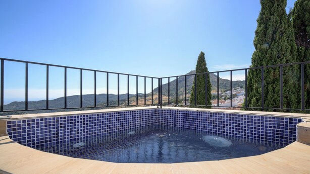 Un bain à remous dont l’intérieur est revêtu de carreaux, entouré d’une terrasse en bois et d’une rambarde en métal, offrant une vue sur un paysage montagneux.
