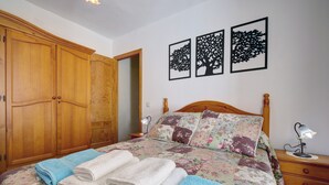 Une chambre avec une armoire en bois, un lit recouvert d’une literie à motifs floraux et trois tableaux représentant des arbres accrochés au mur.