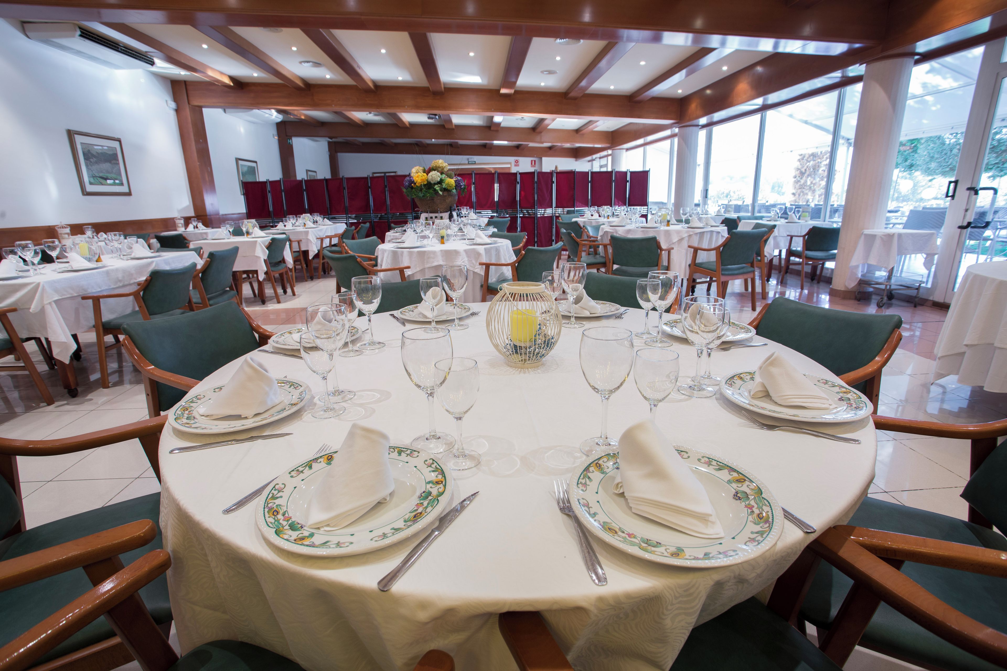 Uma sala de jantar com mesas redondas para quatro pessoas, cada uma com toalhas brancas, cadeiras verdes e louça, copos e talheres dispostos com cuidado.