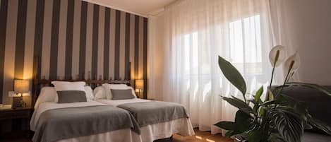 Quarto de hotel com duas camas, uma janela ampla com cortinas leves, piso de madeira e uma planta em vaso sobre uma mesa.