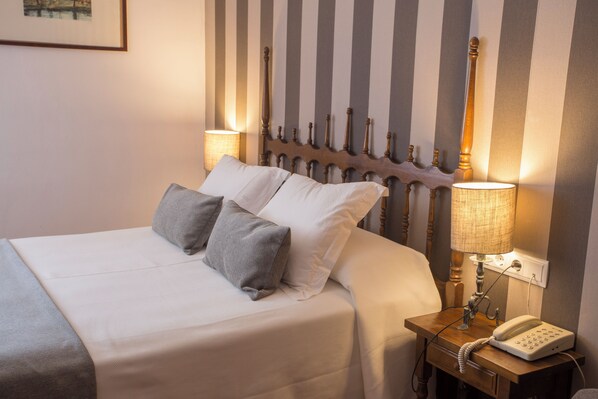 Una cama bien hecha con ropa de cama blanca y gris, un cabecero de madera y una mesita de noche con una lámpara y un teléfono.