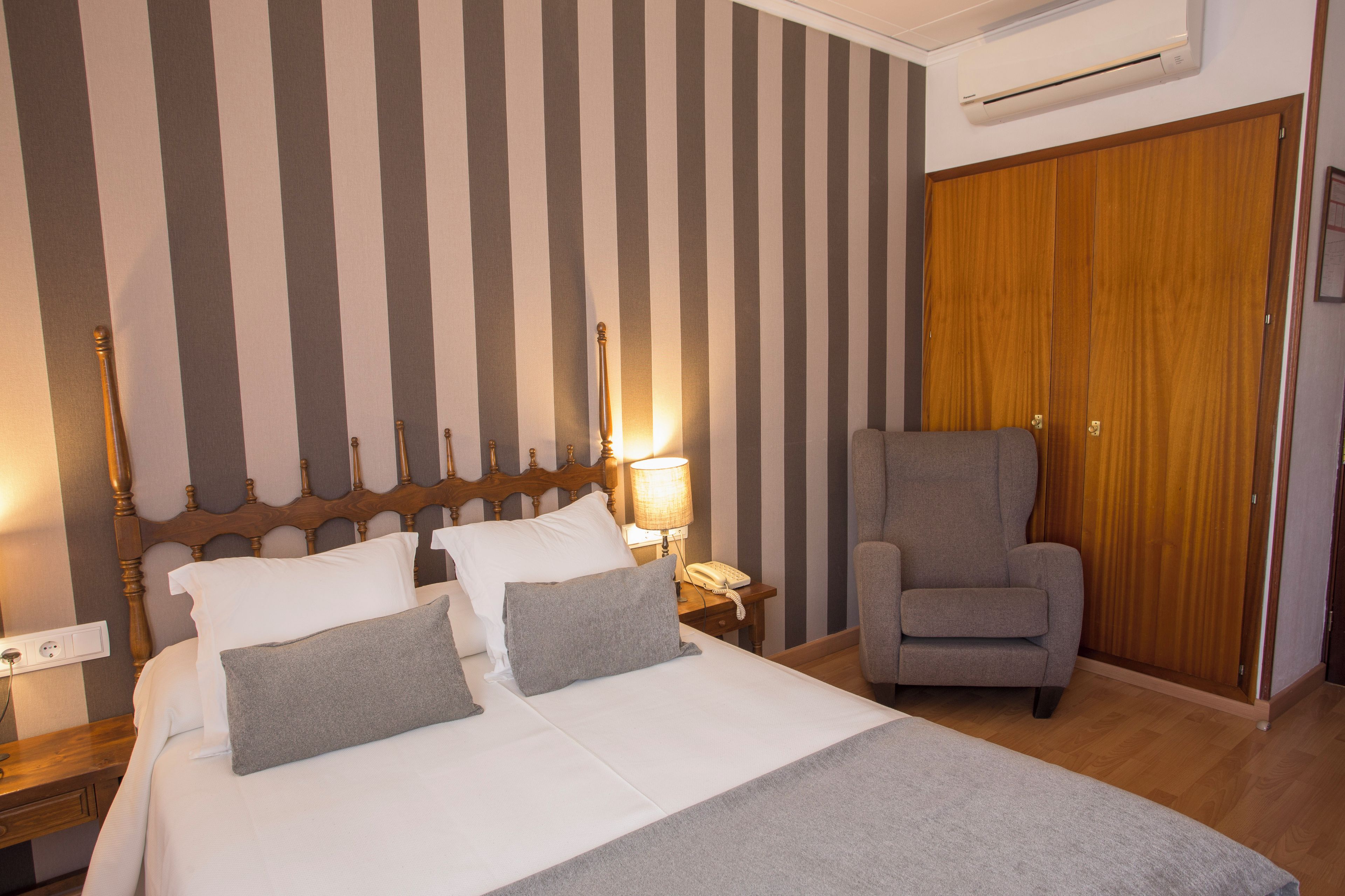 A hotel room with a bed, a chair, a nightstand, a lamp, and a wardrobe.